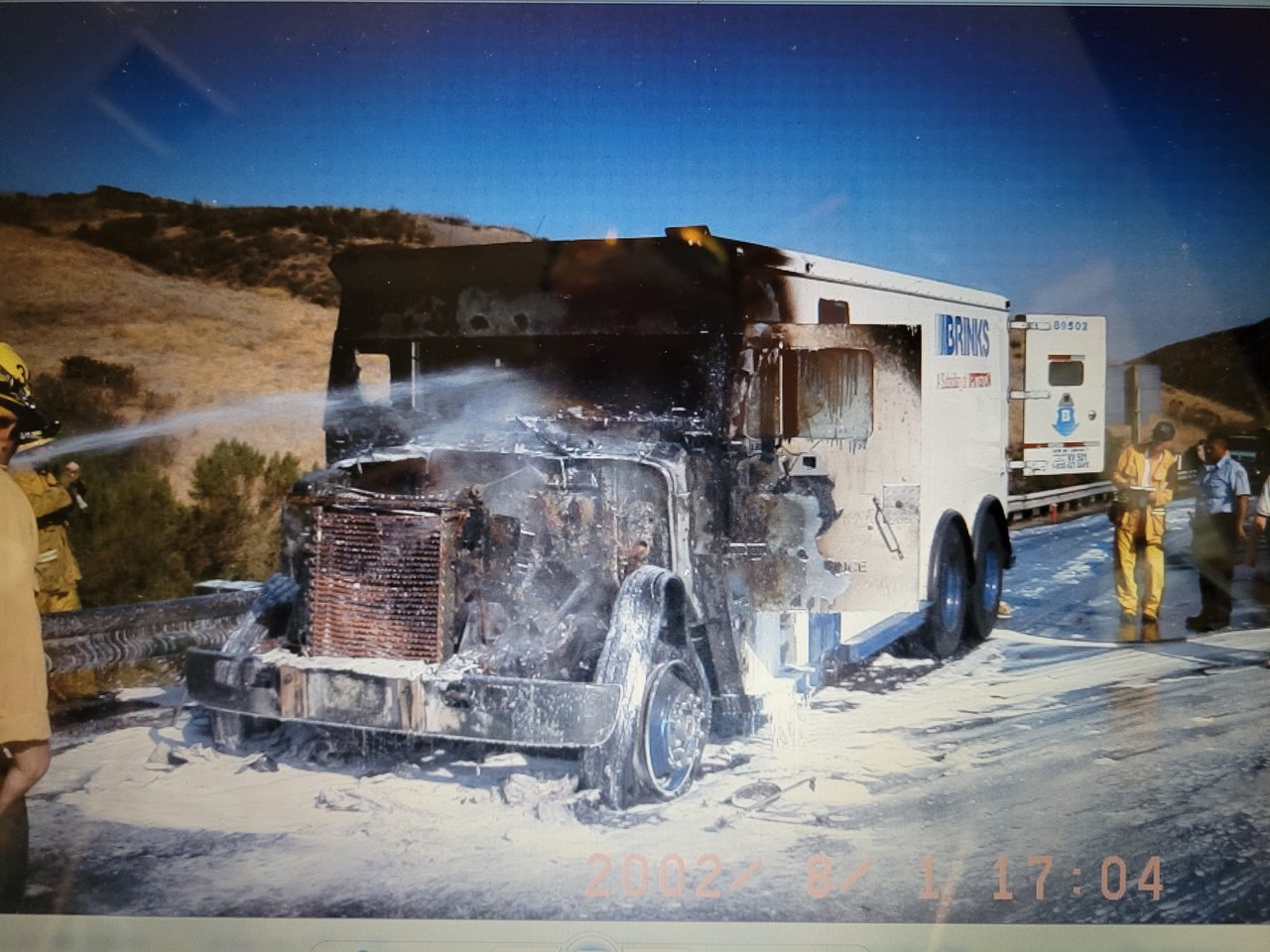 Firefighters applying foam to a fully involved armored transport truck on a rural highway, August 2002